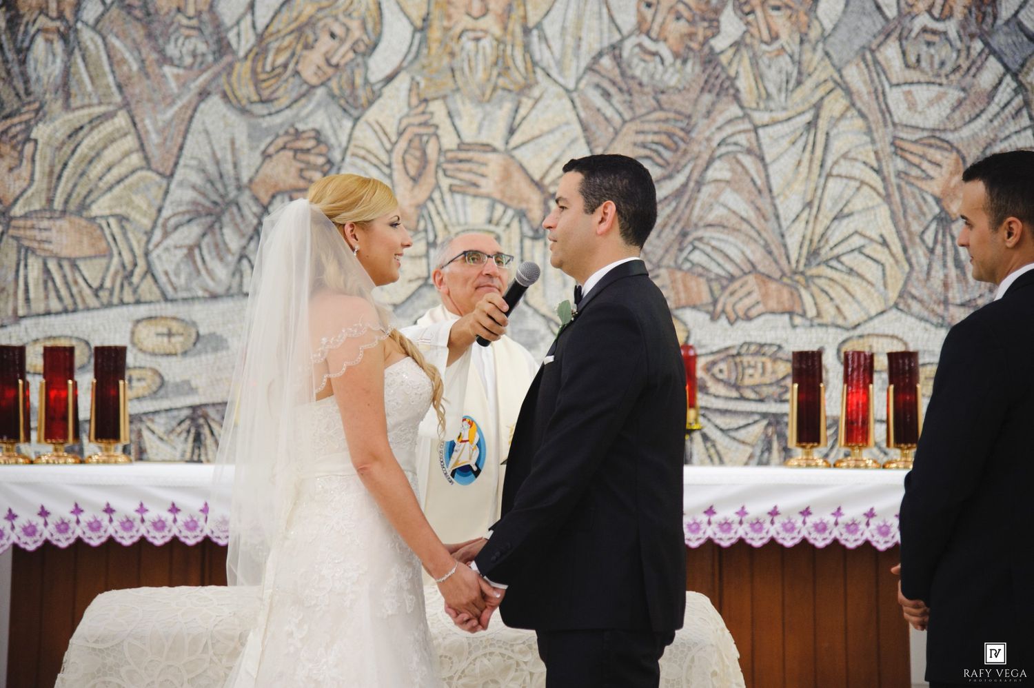 Bride and groom exchanging vows at a Catholic church wedding ceremony with a mosaic mural backdrop.