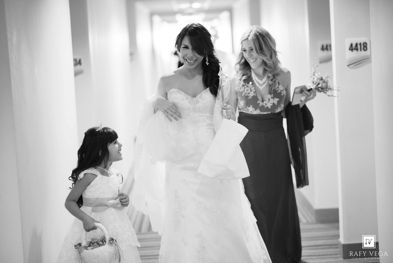 Black and white photo of a bride in a strapless gown walking with a flower girl and bridesmaid in a hotel hallway.