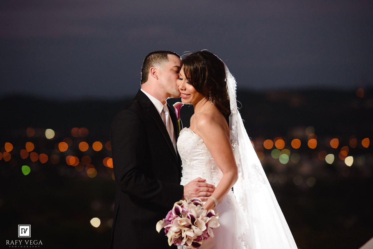 Bride and groom share a tender kiss at night with bokeh city lights glowing behind them.