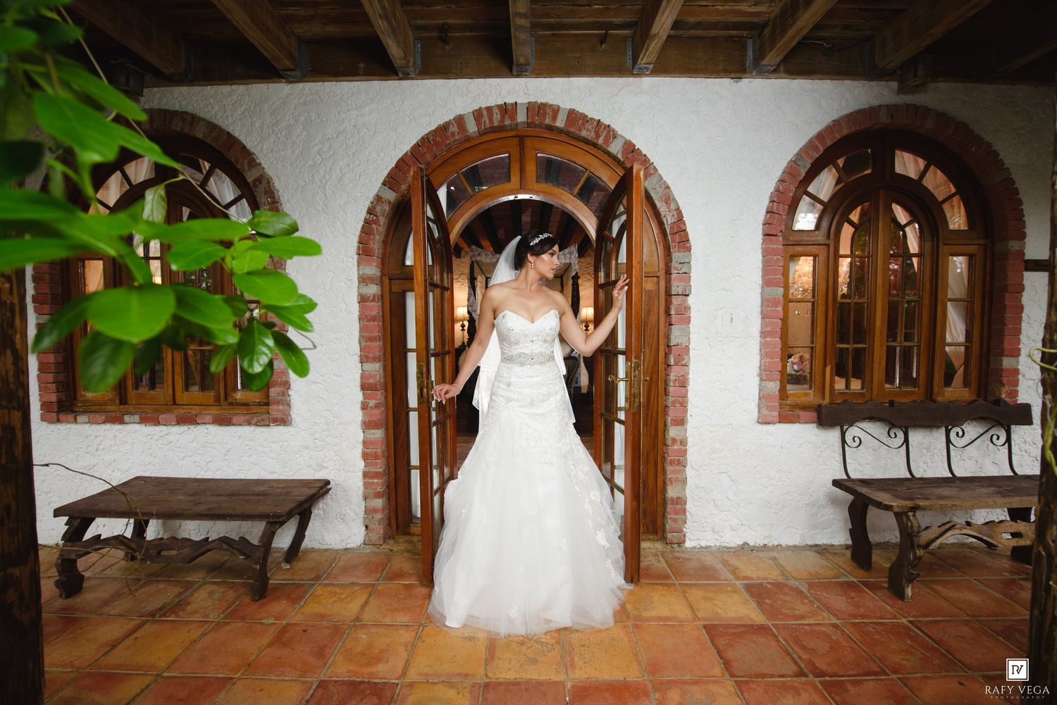 Bride in white wedding gown standing in elegant brick archway doorway at rustic venue with terracotta tile floors.