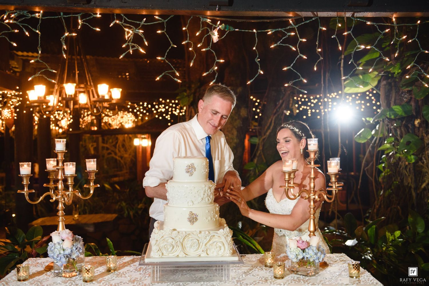 Bride and groom cutting white wedding cake at elegant reception with candleholders and twinkling fairy lights backdrop.