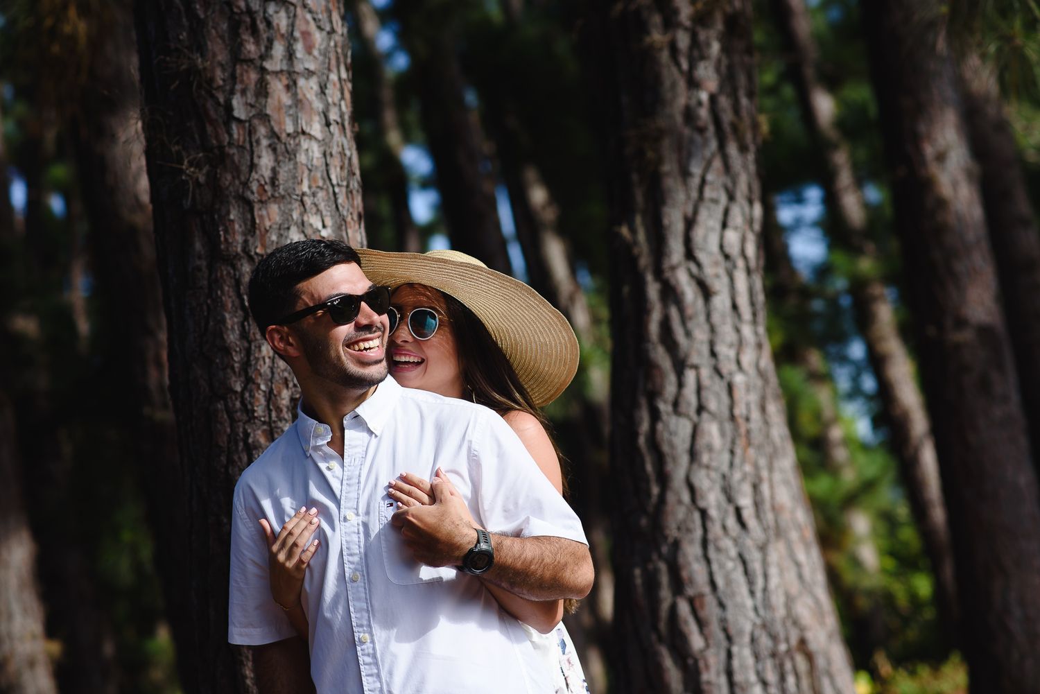Couple in white outfits and sunglasses embracing by pine trees, woman wearing straw hat, outdoor forest setting.