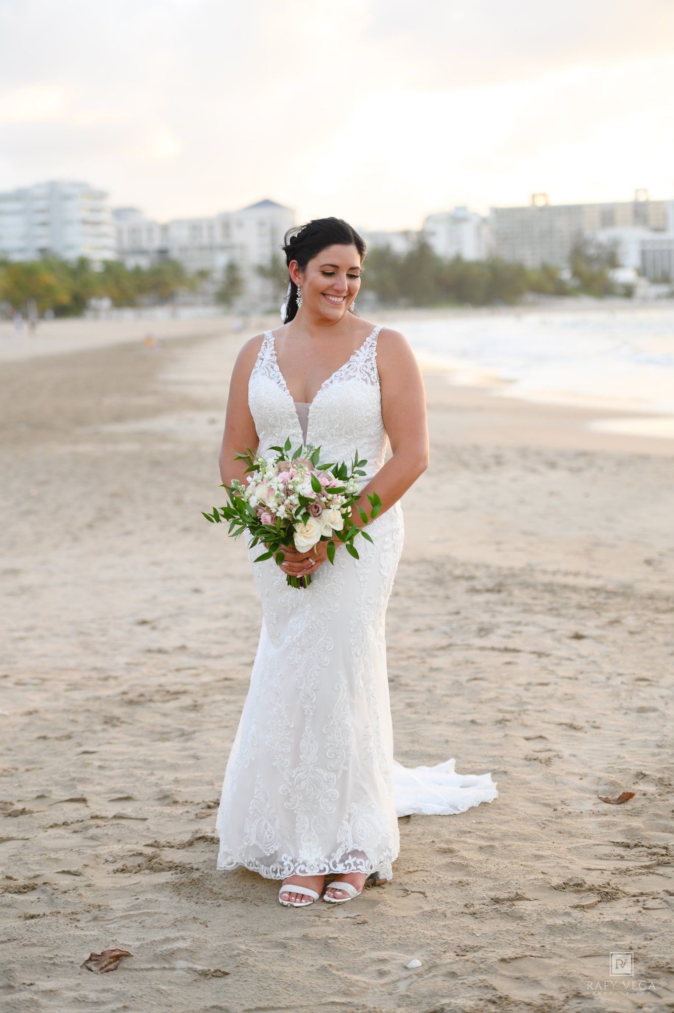 Bride in white lace mermaid gown holding floral bouquet on sandy beach with ocean and resort backdrop.