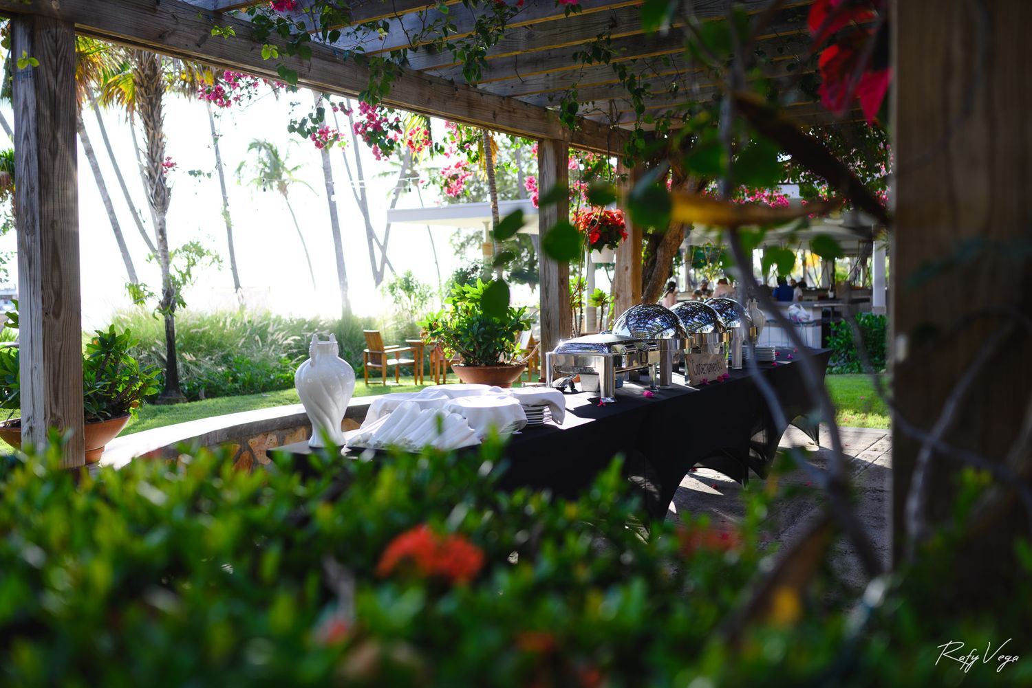Elegant outdoor restaurant patio with lush tropical flowers, set tables, and ocean views under a wooden pergola.