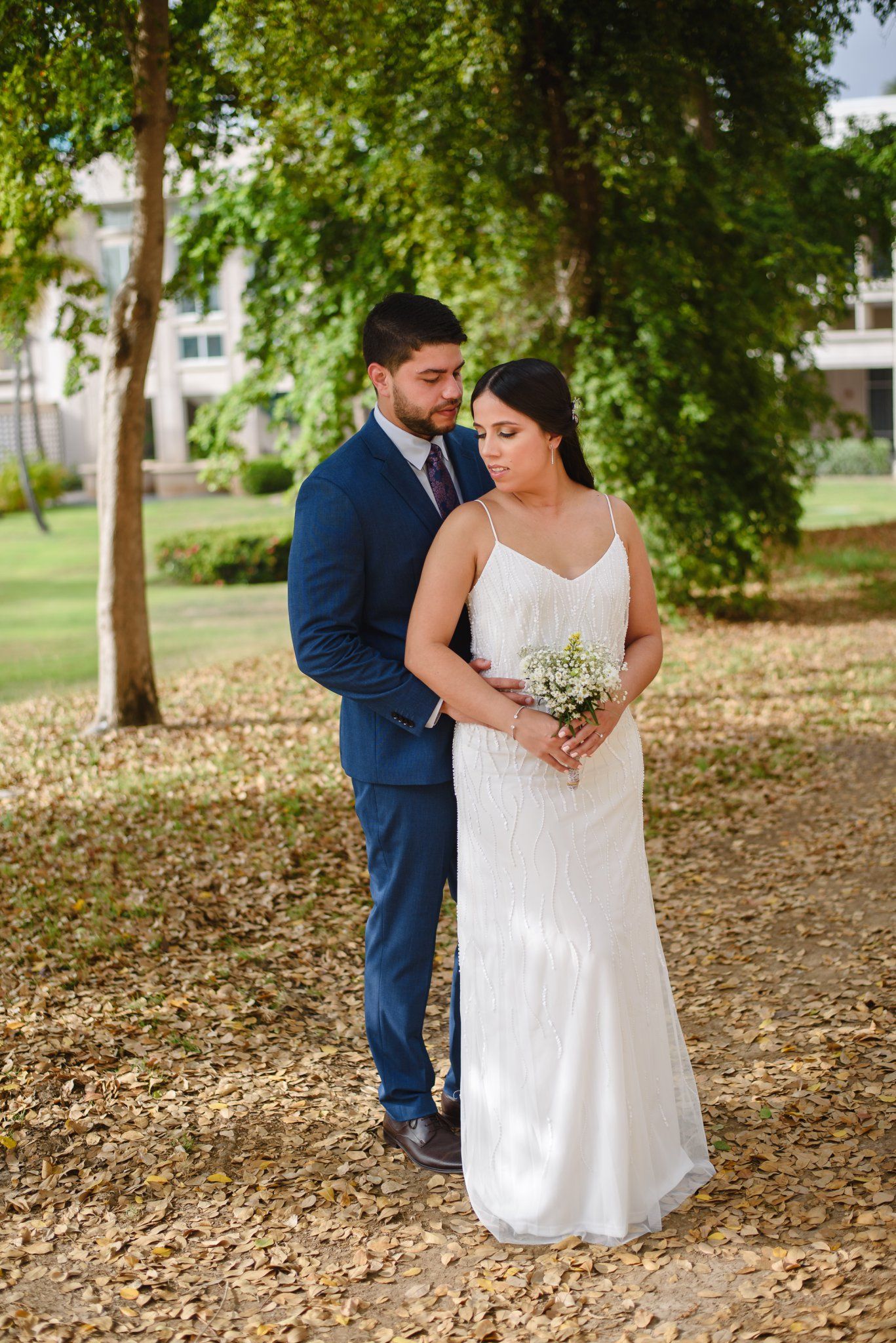 Bride in white gown holding bouquet stands with groom in navy suit outdoors among trees on fallen leaves.