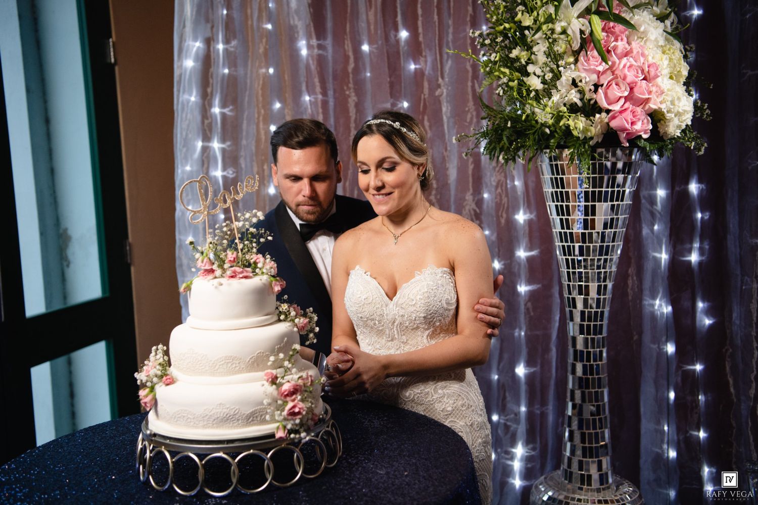 Bride and groom cutting their white three-tier wedding cake at elegant reception with fairy lights and floral arrangements.
