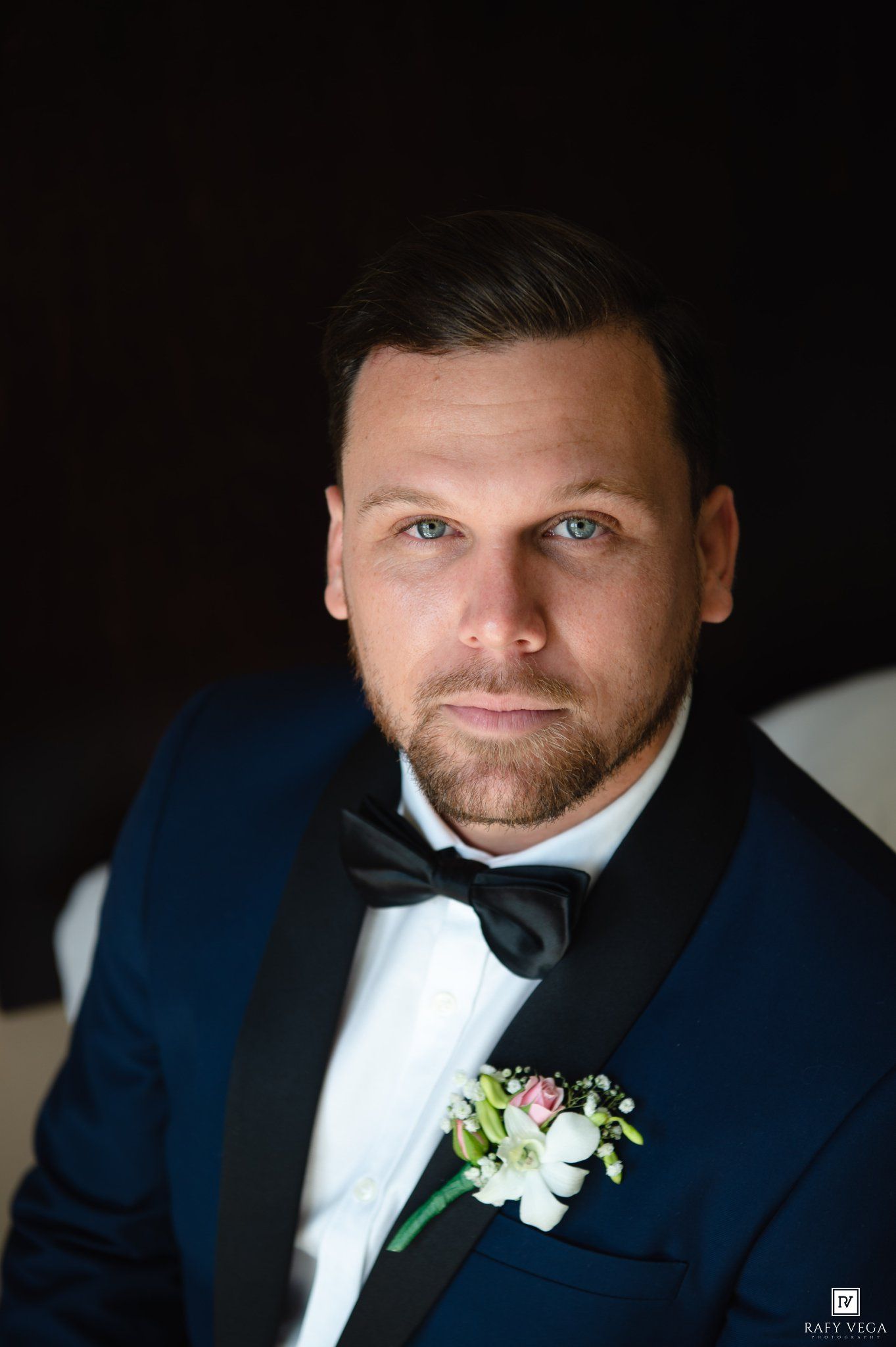Groom in navy blue tuxedo with black bow tie and floral boutonniere, posing for a formal wedding portrait.