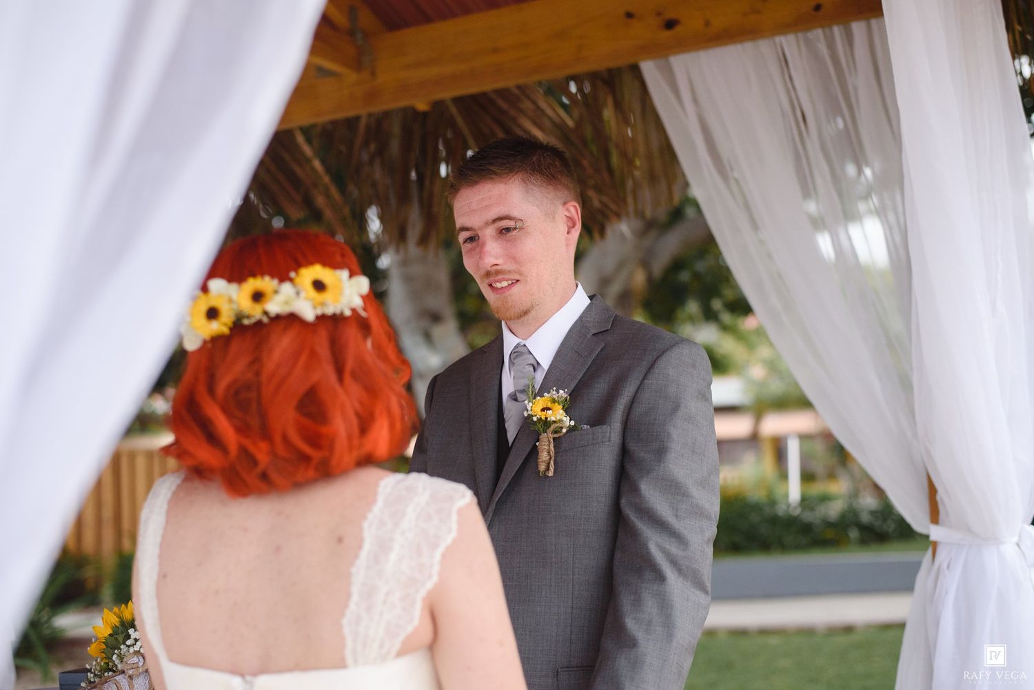Groom in gray suit smiles at red-haired bride wearing flower crown during outdoor wedding ceremony.