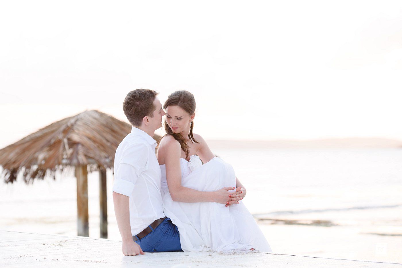 Couple sitting on beach, pregnant woman in white dress, man in white shirt and jeans, thatched umbrella, serene ocean backdrop.
