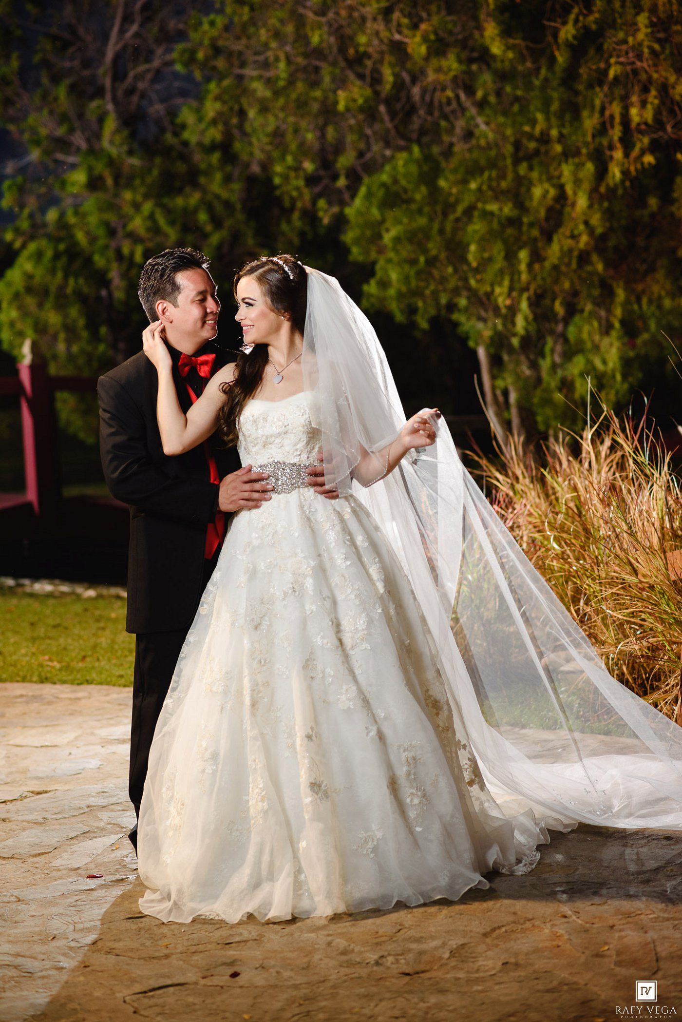 Bride in white lace ball gown with veil and groom in black suit share a romantic moment outdoors at night.