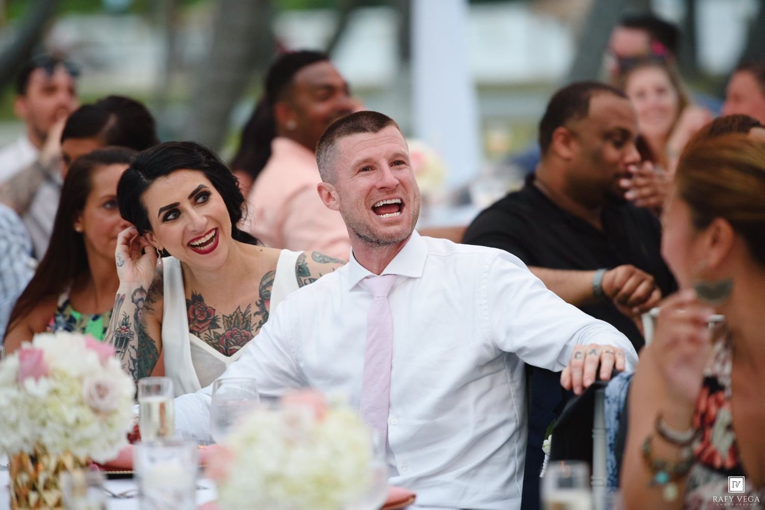 Guests laughing and smiling at a wedding reception, seated at a decorated table outdoors.