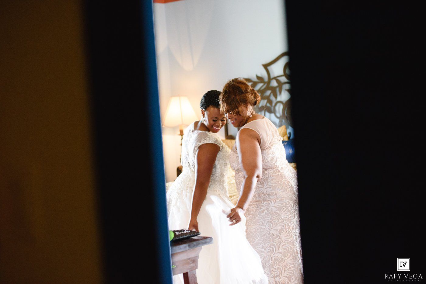 Two women in elegant white gowns prepare for a wedding ceremony in a softly lit room.