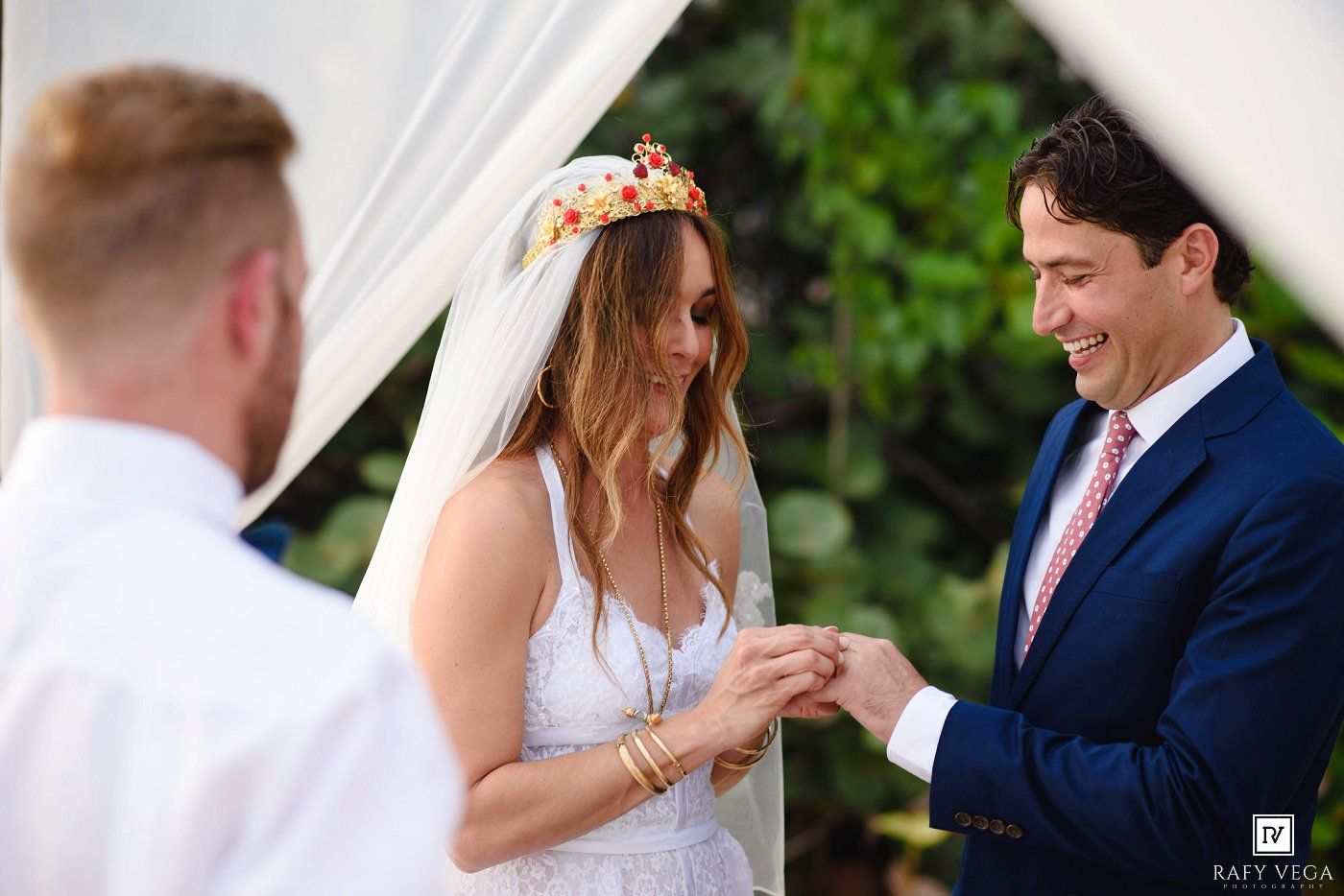 Bride and groom exchanging rings during outdoor wedding ceremony, bride wearing floral crown and veil, groom in navy suit.