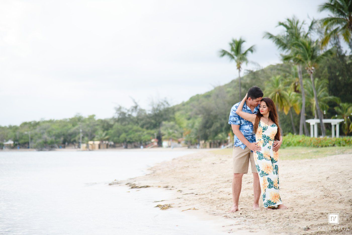 Couple embracing on a tropical beach with palm trees and calm waters in the background.