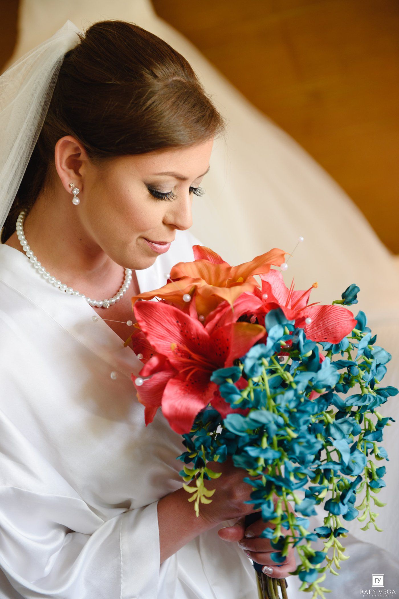 Bride in white gown and veil holds vibrant bouquet of red lilies and teal flowers, wearing pearl jewelry.