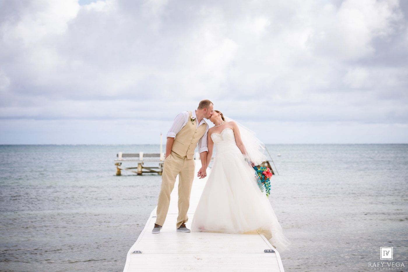 Bride and groom share a romantic kiss on a white pier overlooking a calm turquoise ocean on their wedding day.