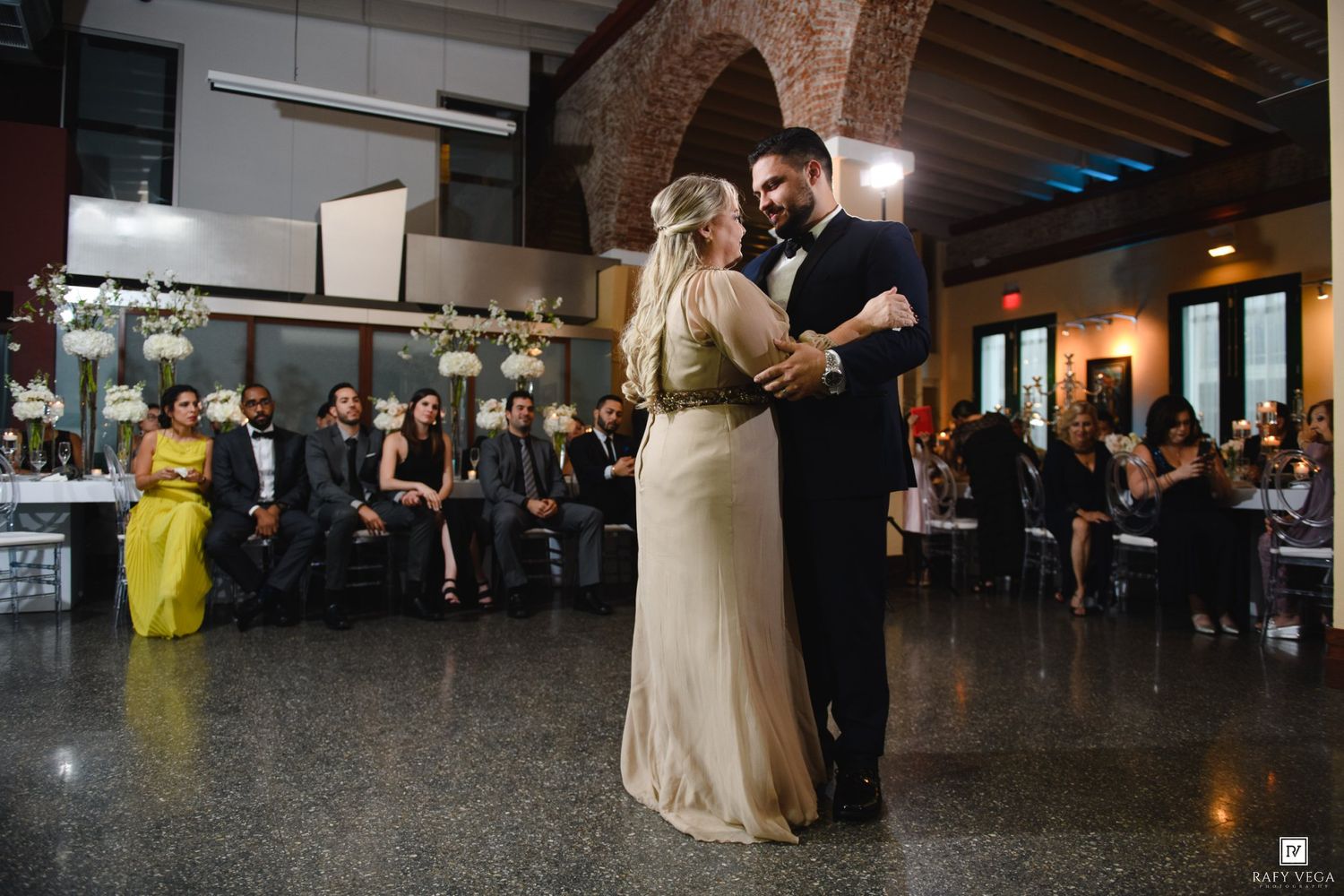 A couple shares their first dance at an elegant wedding reception with guests seated in the background.