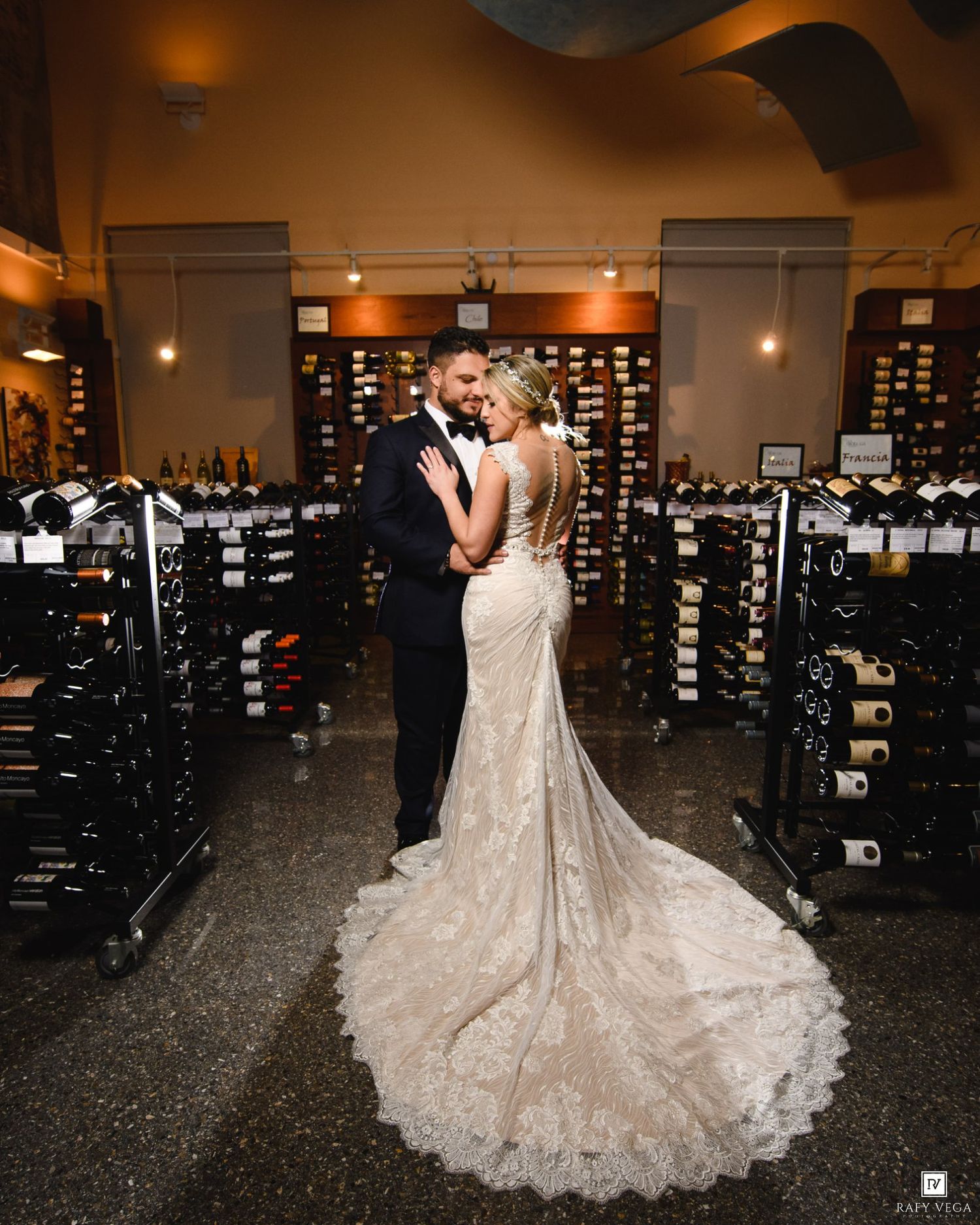 Bride and groom share romantic embrace in elegant wine cellar, bride wearing lace mermaid gown with long train.