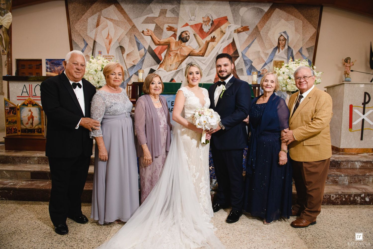 Wedding party poses in church with bride in white gown and groom in suit surrounded by family members.