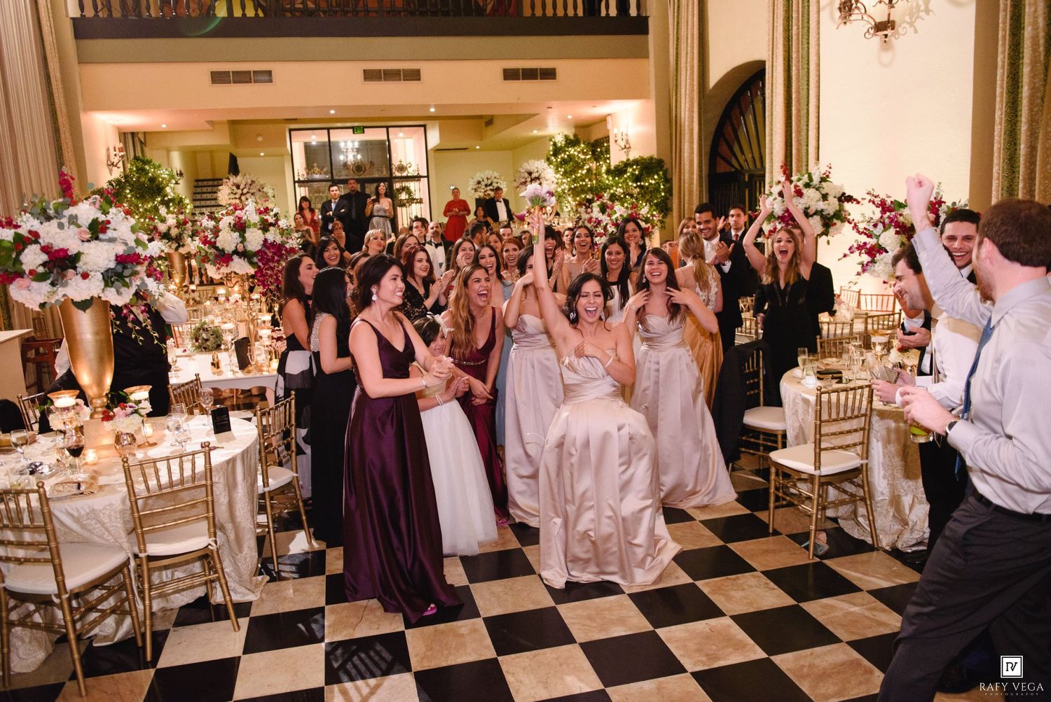 Bride surrounded by bridesmaids in purple gowns celebrating at elegant wedding reception on black and white checkered floor.
