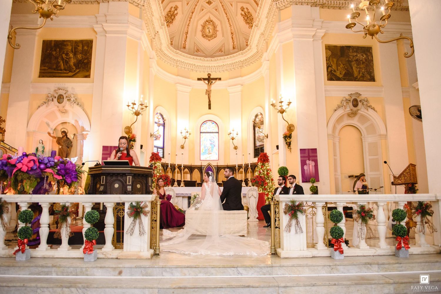 Elegant Catholic church wedding ceremony with floral decorations, bride and groom at ornate white altar with cross.