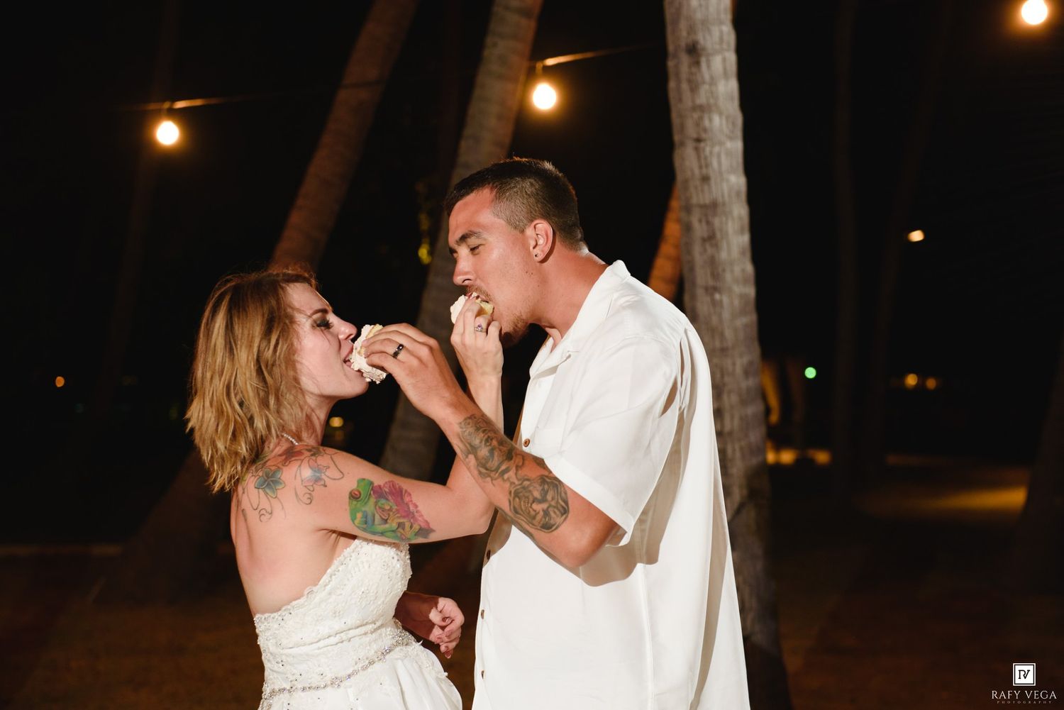 Couple in white outfits sharing cake at night outdoor wedding celebration with palm trees and string lights.