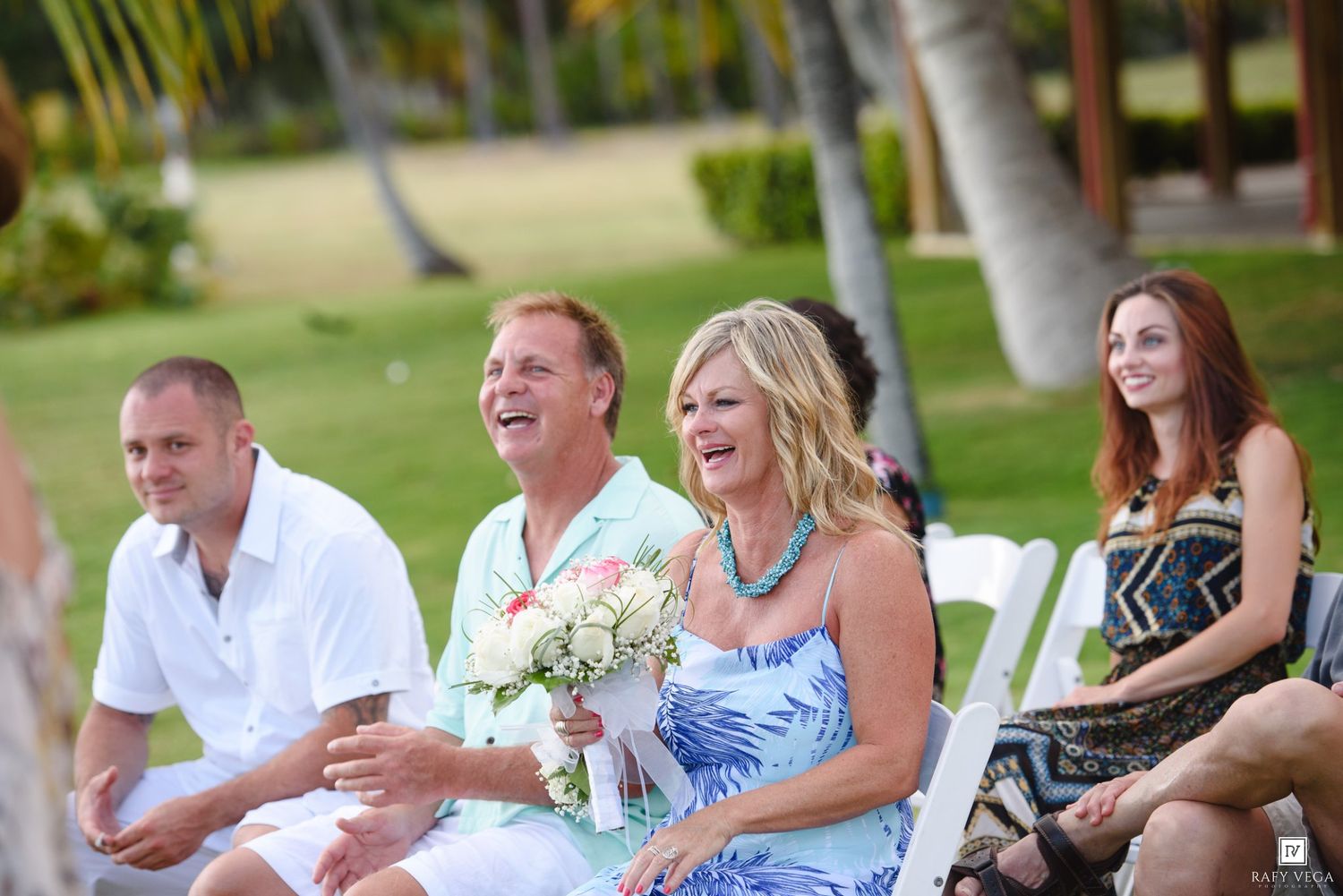 Guests laughing and smiling at an outdoor tropical wedding ceremony, woman holding a coral bouquet.