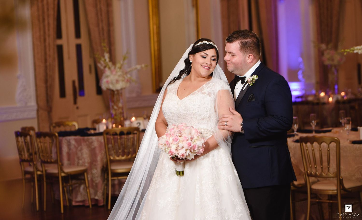 Bride and groom share a romantic moment in an elegant ballroom, bride holding a pink bouquet in a white gown.