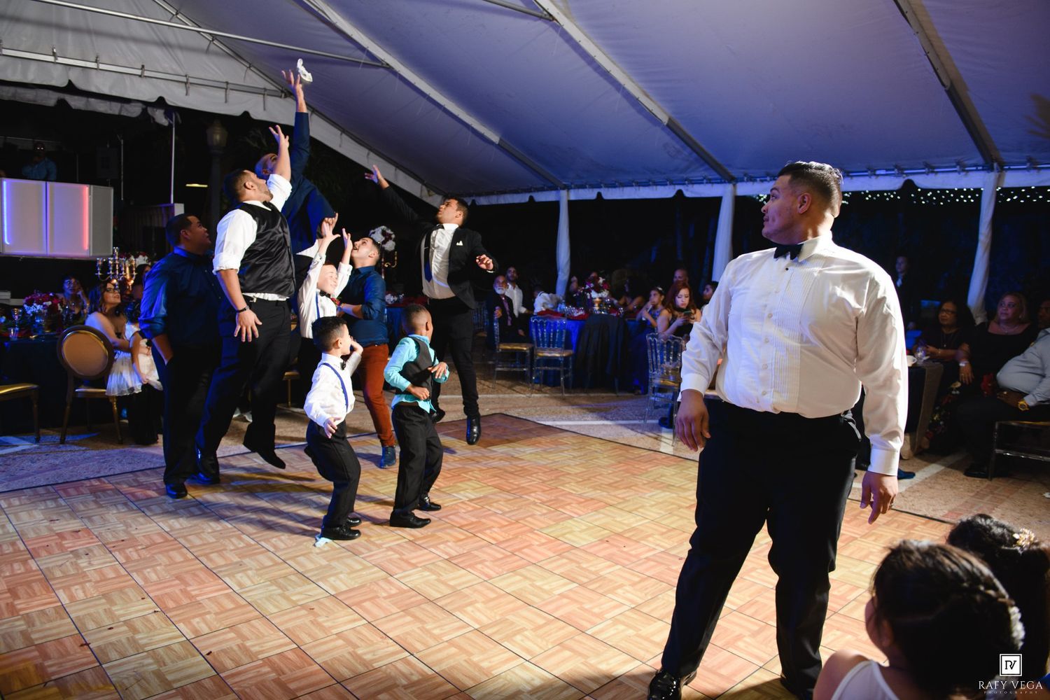 Men and boys dancing energetically at an elegant outdoor wedding reception under a white tent.
