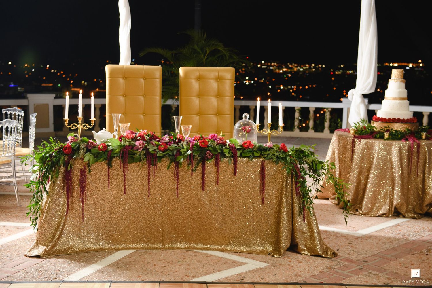 Elegant gold sequin sweetheart table with floral garland and tall chairs at an outdoor night wedding reception.