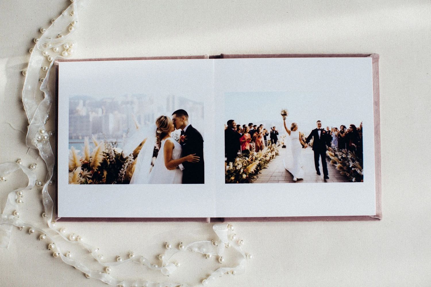 Open wedding photo album displaying bride and groom portraits and outdoor ceremony aisle photos on white linen background.