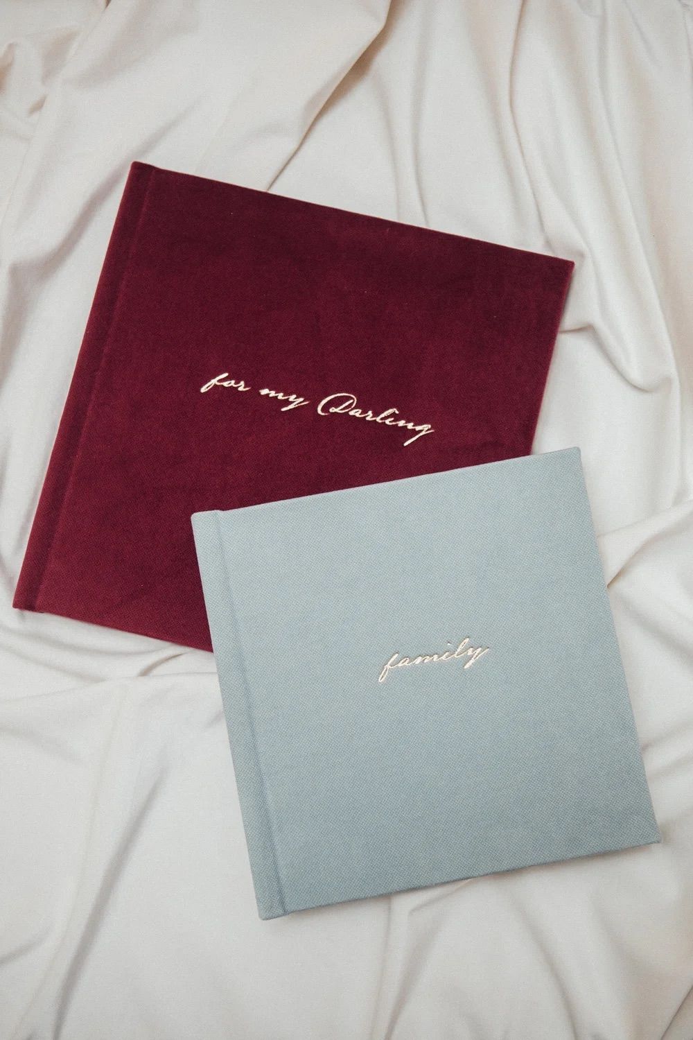 Two elegant hardcover journals on white fabric, one burgundy velvet reading 
