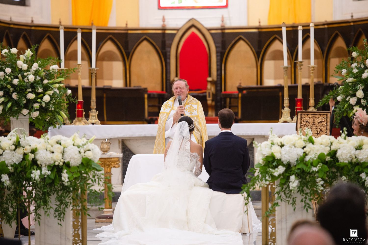 Priest officiating a wedding ceremony at a church altar adorned with white floral arrangements and candles.