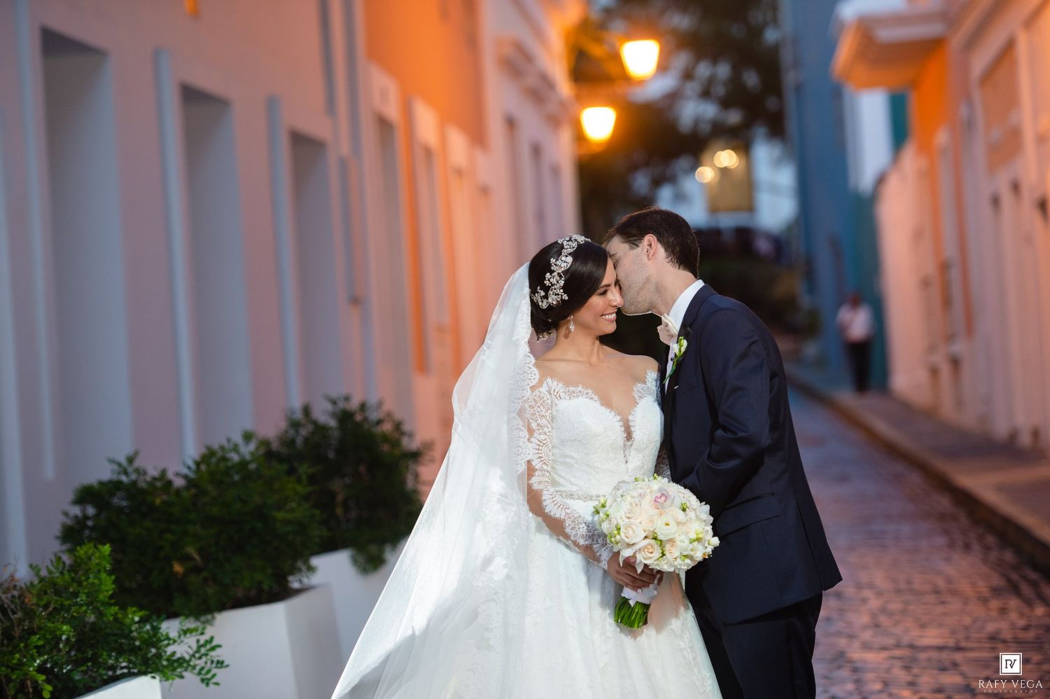 Bride and groom share a romantic kiss on a cobblestone street, bride in a white ball gown holding a bouquet.