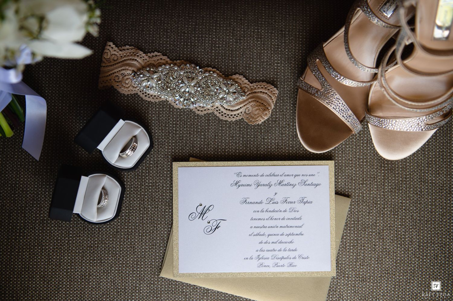Elegant wedding flat lay featuring invitation, diamond rings, silver heels, rhinestone belt, and navy ribbon on brown surface.