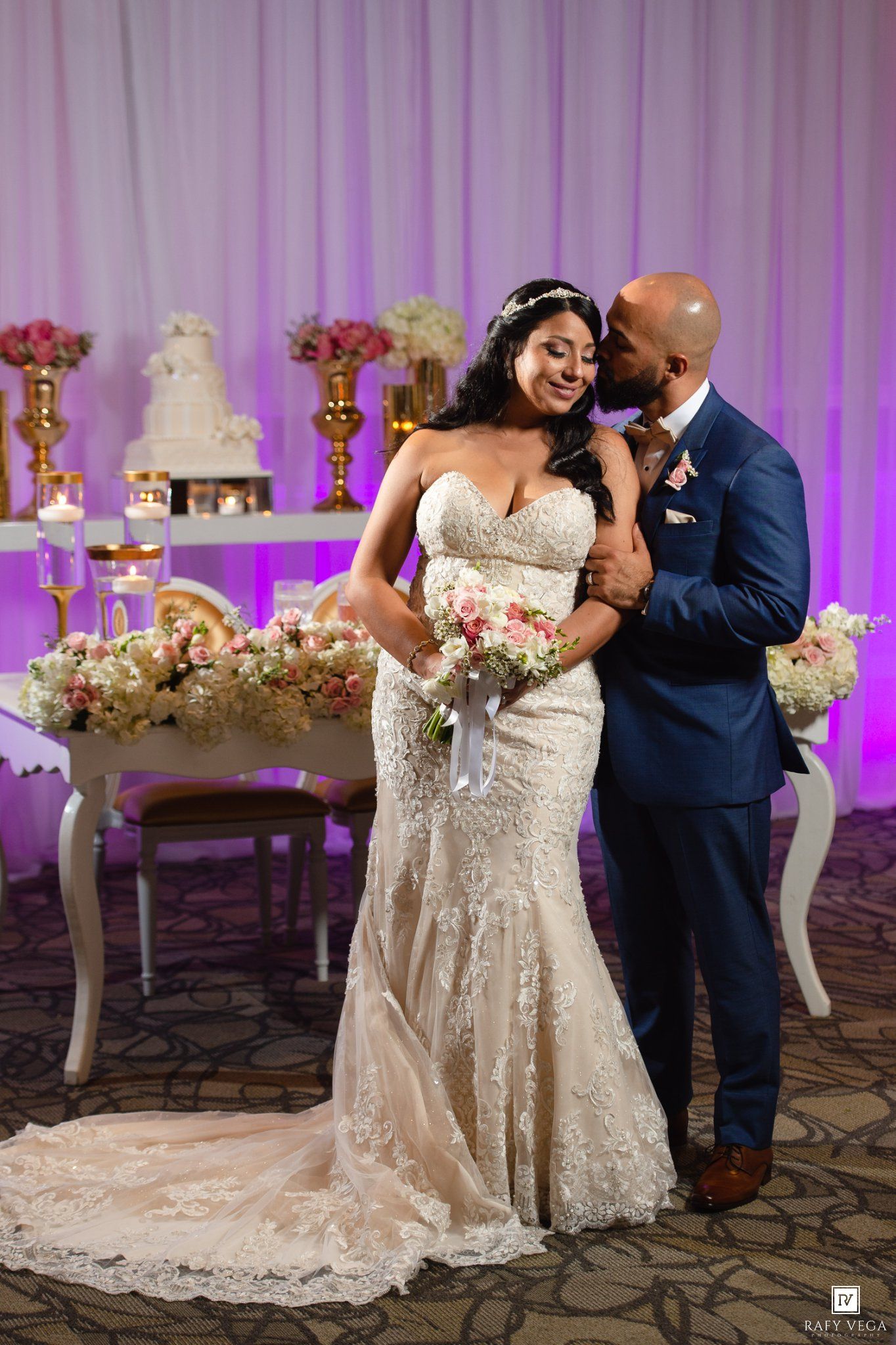 Bride in lace mermaid gown holds bouquet as groom in navy suit whispers in her ear at elegant wedding reception.