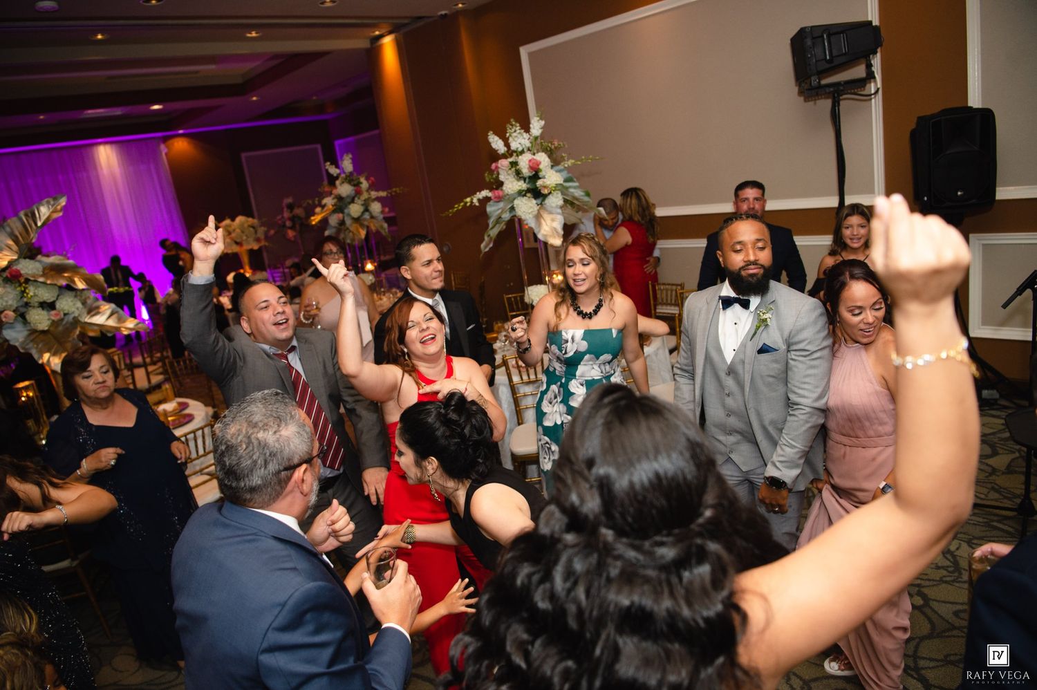 Guests celebrate and dance together at an elegant wedding reception with floral arrangements and colorful formal attire.
