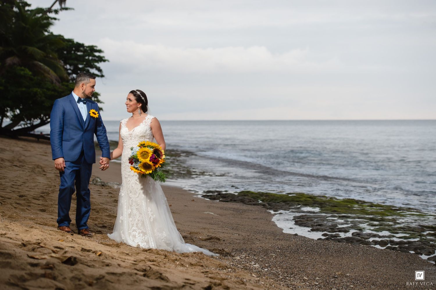 Bride and groom holding hands on a sandy beach shoreline, bride holding sunflower bouquet in white lace gown.
