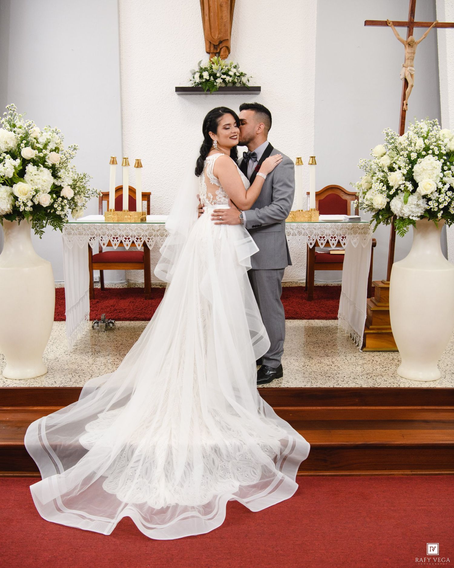 Bride in stunning white gown with long veil kisses groom in gray suit at church altar.