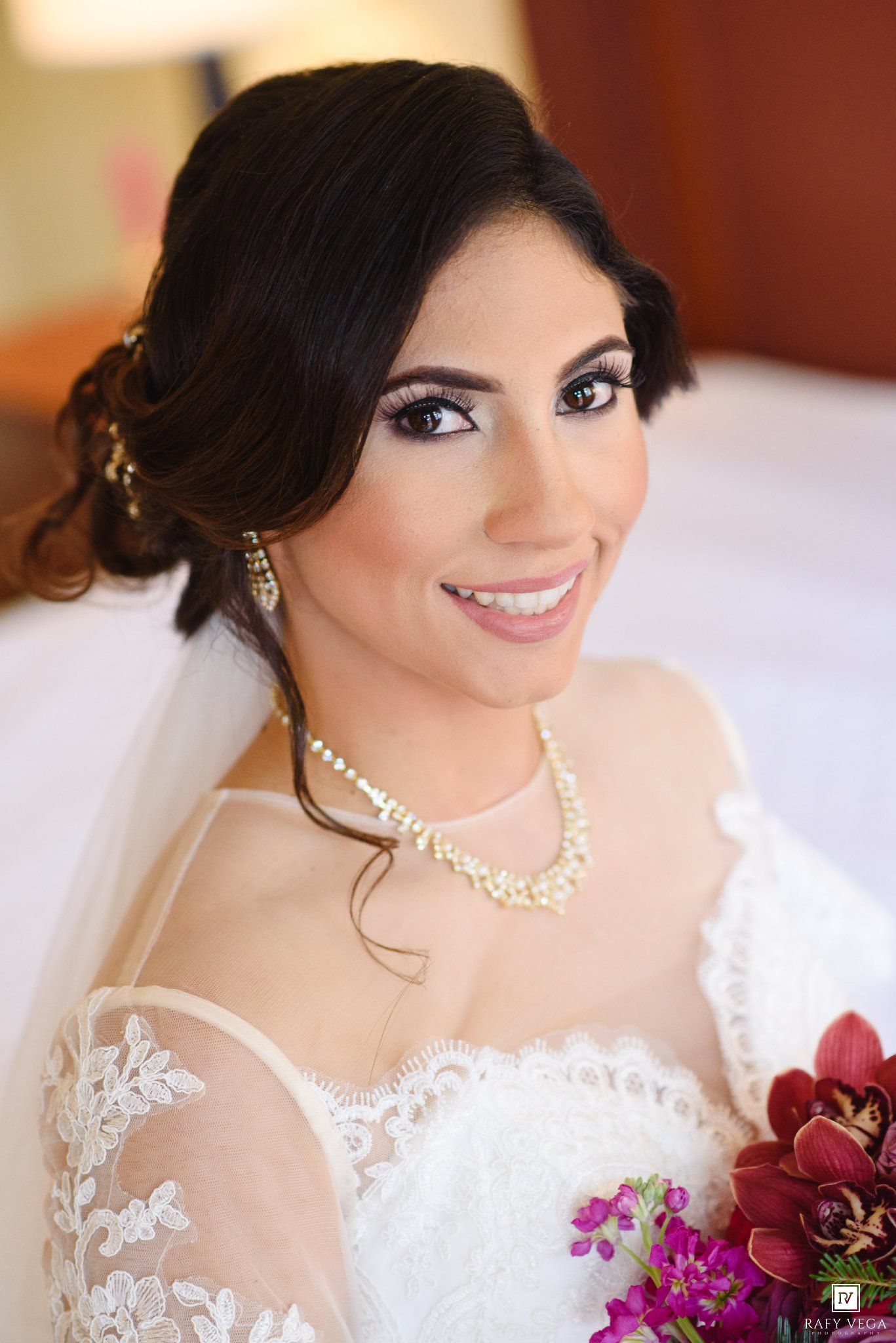 Elegant bride in white lace gown with pearl necklace, veil, and bridal makeup holding a bouquet.