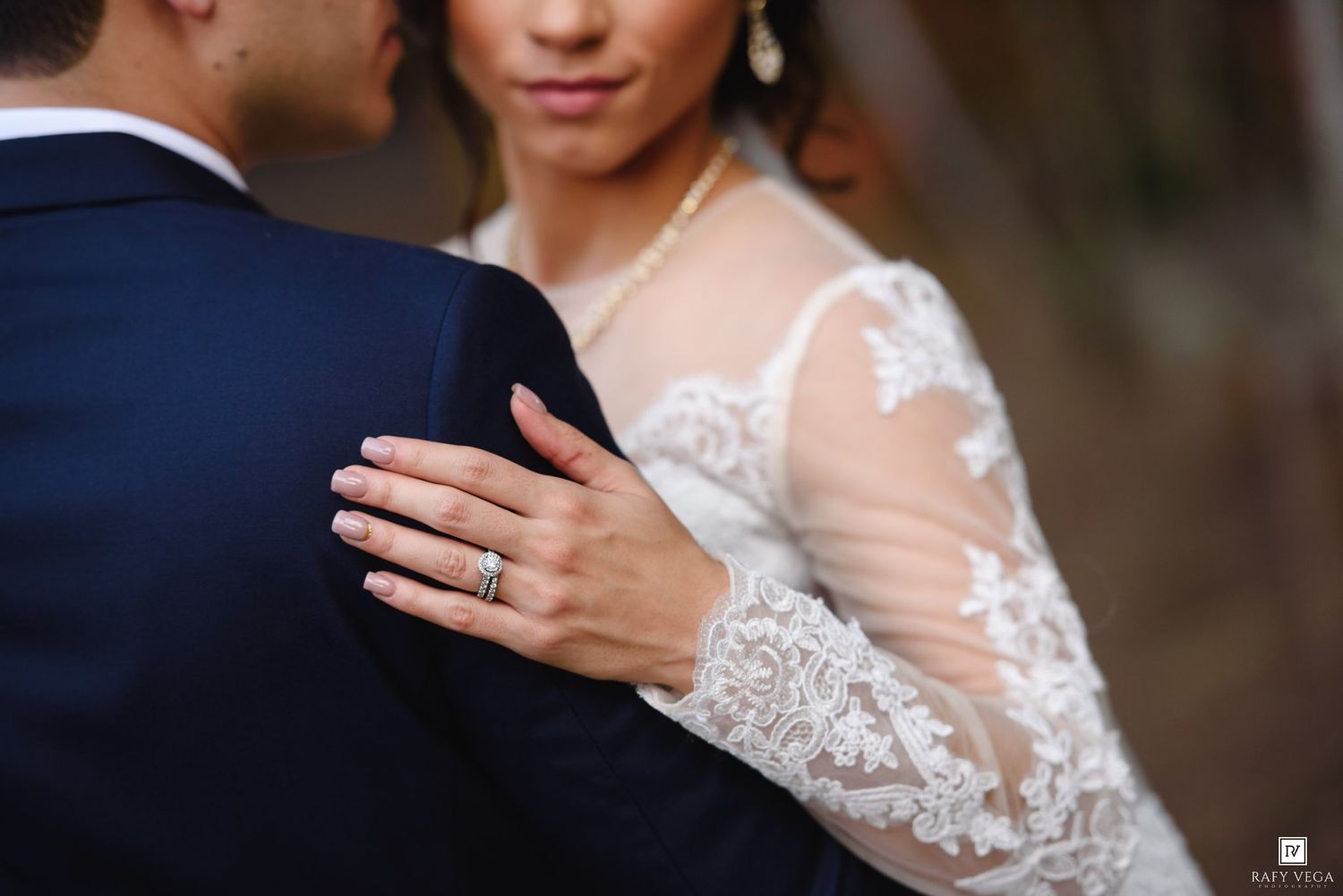 Bride in lace wedding gown with diamond ring rests hand on groom's navy suit jacket during wedding ceremony.