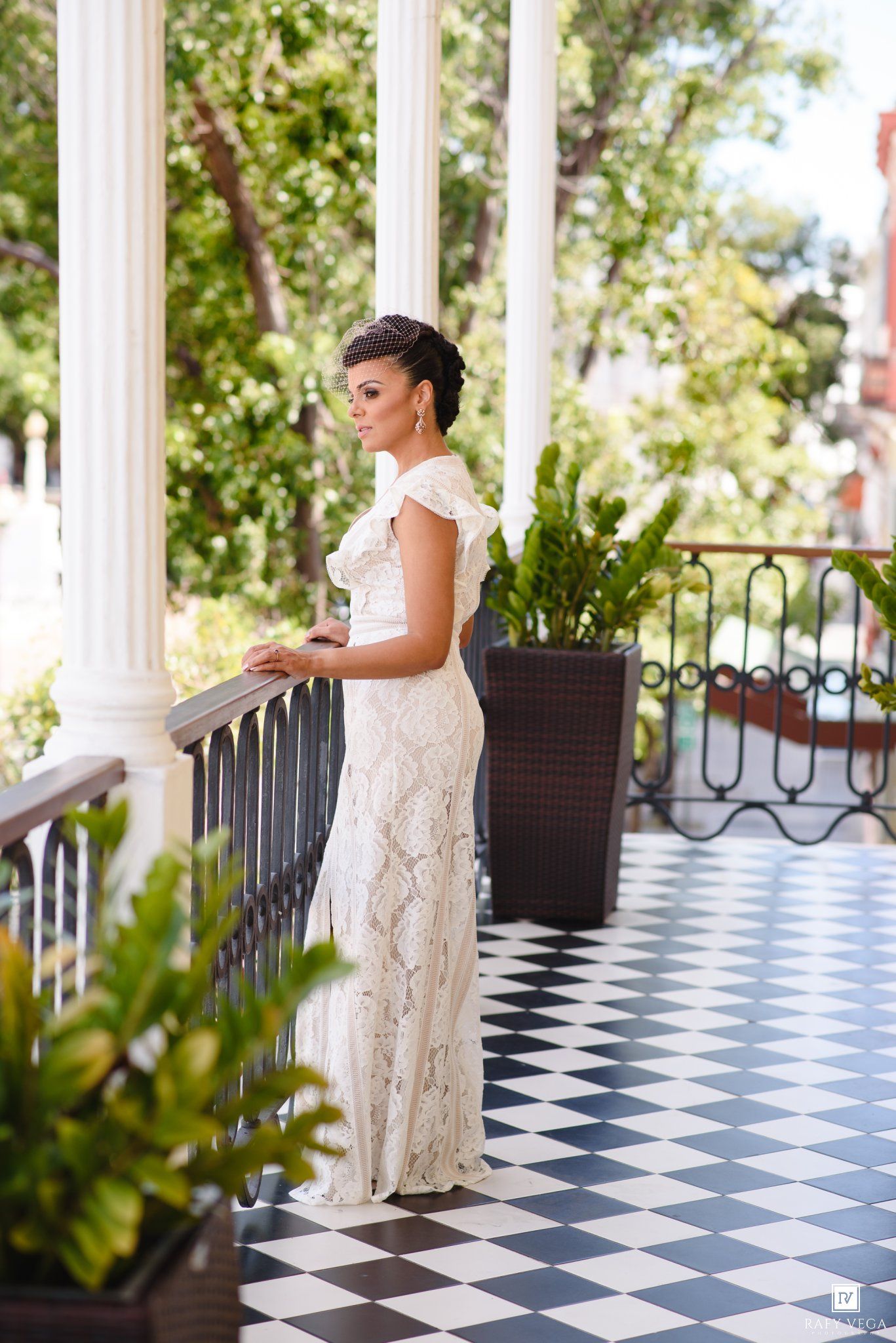 Elegant bride in white lace gown stands on a checkered balcony with columns and lush greenery.