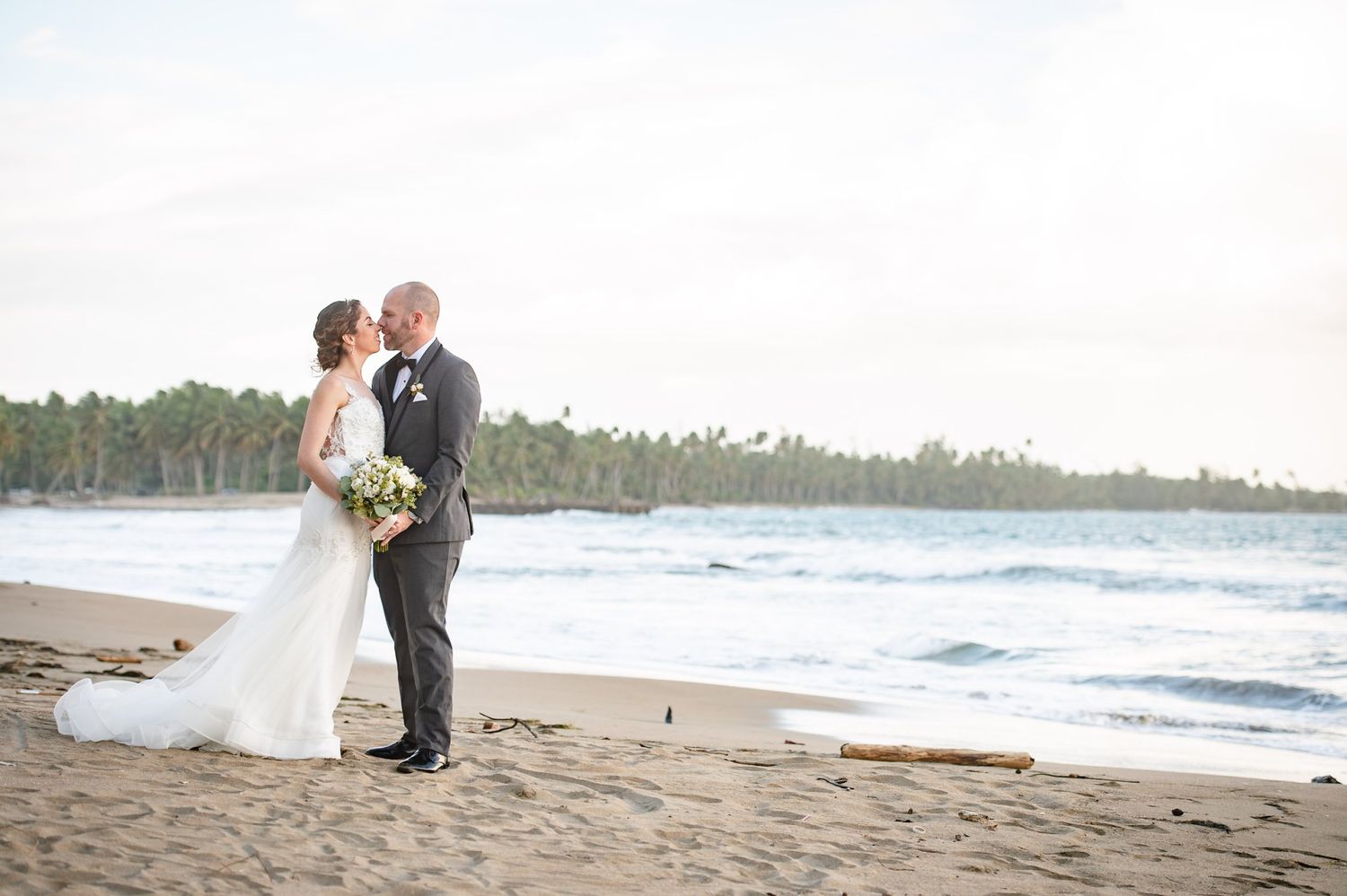 Bride and groom share a romantic kiss on a sandy beach with ocean waves and tropical trees in the background.