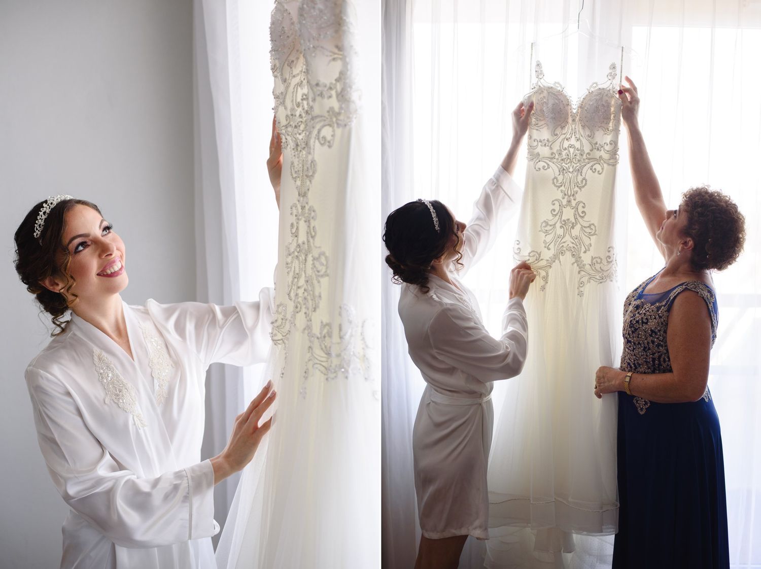 Bride in white robe smiles at her lace wedding gown as bridesmaids help hang it near a bright window.