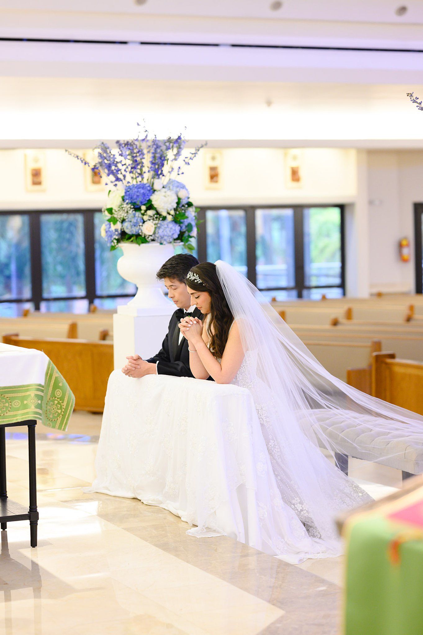 Bride and groom kneel together in prayer at a church altar adorned with blue hydrangea floral arrangements.