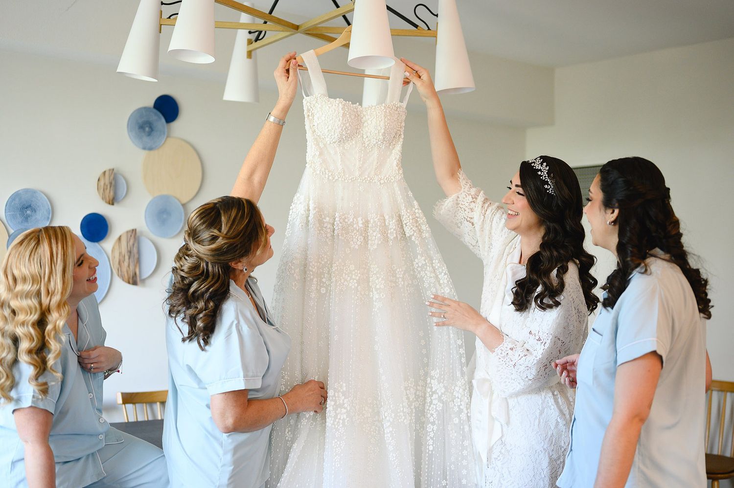 Bride and bridesmaids in blue robes admiring a lace wedding dress hanging on a gold chandelier.