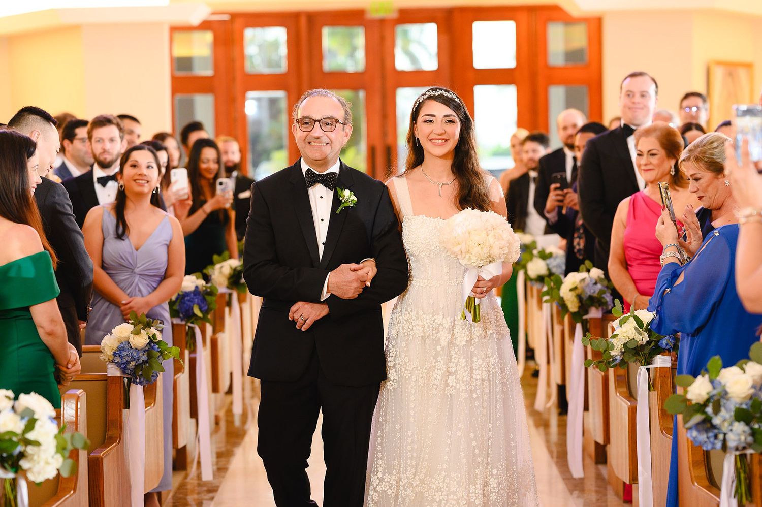 Bride walks down church aisle with escort, wearing lace gown and tiara, guests seated in decorated pews.