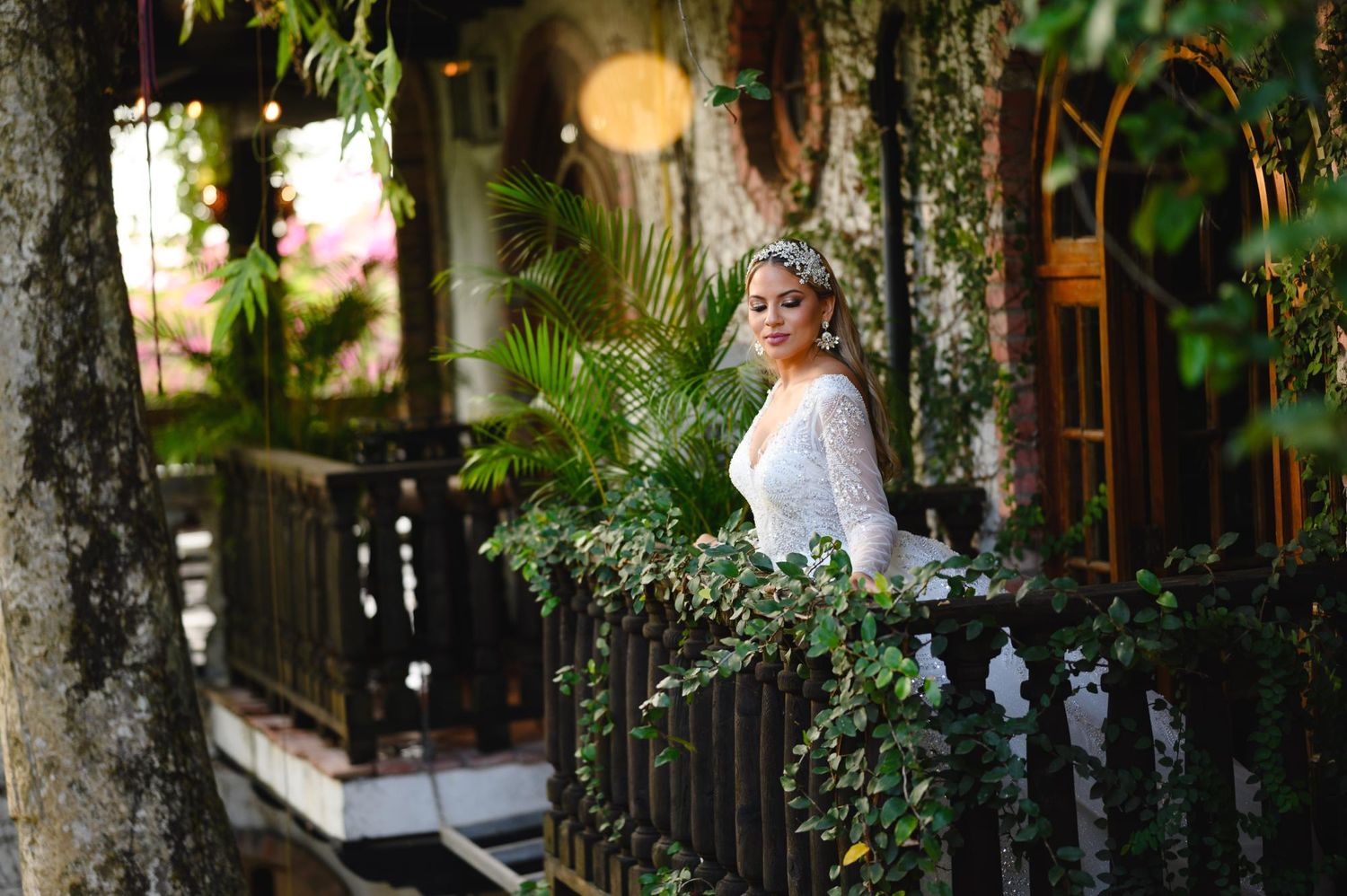 Bride in white lace gown stands on ivy-covered balcony surrounded by lush tropical greenery.