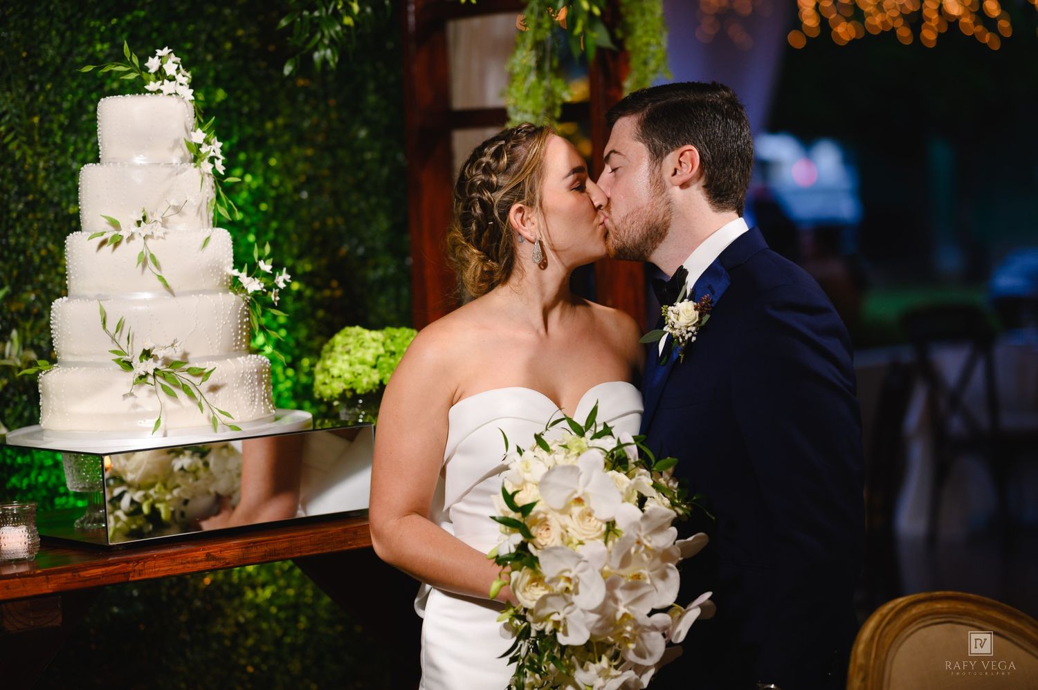 Bride and groom share a kiss beside a white floral wedding cake at an elegant indoor reception venue.