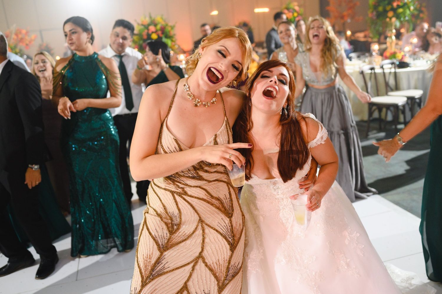 Two women laughing and dancing joyfully on the wedding reception dance floor in formal attire.